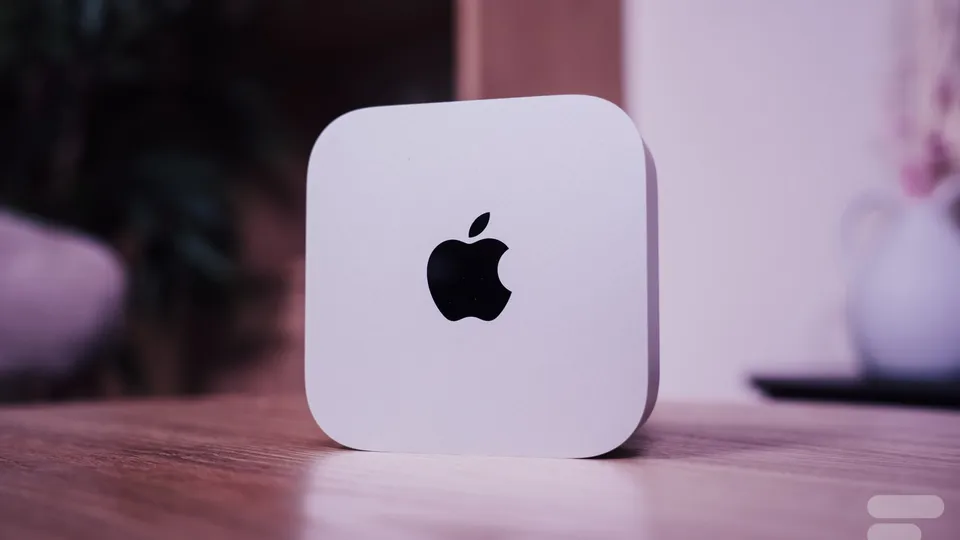 Mac mini M6 gris clair au logo Apple noir, posé sur un bureau en bois devant un fond rose flou